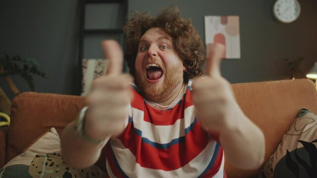 Young ecstatic man shouting with excitement, clenching fists, giving thumbs up and celebrating goal while watching sports game on couch in living room. POV, whip zoom shot