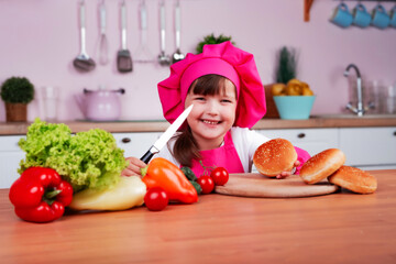Funny happy little beautiful girl in a chef costume prepares burgers while sitting at a table in the kitchen. Healthy eating.	