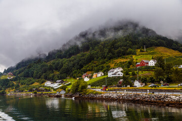 Fototapeta premium Scenic Coastal Village with Lush Green Hills and Misty Mountains Under Overcast Skies - A Serene View of Norway's Natural Beauty Along the Water's Edge