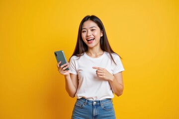 Naklejka premium Cheerful Young Asian Woman in Casual White T-shirt Holding a Smartphone while Smiling Against a Bright Yellow Background