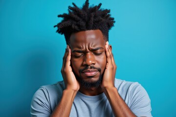 Obraz premium A Young African American Man with Natural Hair Holding His Head in Distress Against a Blue Background, Reflecting Stress or Overwhelm