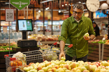 Portrait of mature Indian man shopping in supermarket and choosing fresh apples in produce aisle filled with ripe fruits, copy space