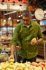 Vertical portrait of adult Indian man choosing fresh apples in produce aisle filled with ripe fruits while shopping in supermarket