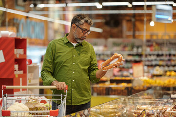 Side view portrait of mature Indian man choosing sausage in meat isle and reading ingredients label while doing grocery shopping in supermarket, copy space