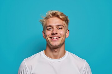 A young Caucasian male with blonde hair smiles brightly against a vibrant turquoise background, exuding positivity and confidence in a casual white t-shirt.