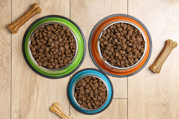 Dry pet food in feeding bowls and chew bones on wooden floor, flat lay
