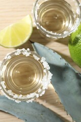 Tequila shots with salt, limes and agave leaves on wooden table, flat lay