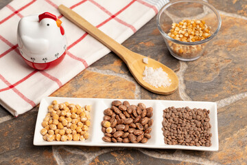 un plato con legumbres, porotos, lentejas, garbanzos, atrás una cuchara de madera con sal gruesa, al fondo un vaso con maiz, un reloj de cocina sobre un paño de cocina 