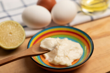 una cuchara de palo con una muestra de mayonesa en primer plano, abajo un colorido plato con mayonesa, al fondo huevos, un trozo de limón, un vaso con aceite y un paño de cocina