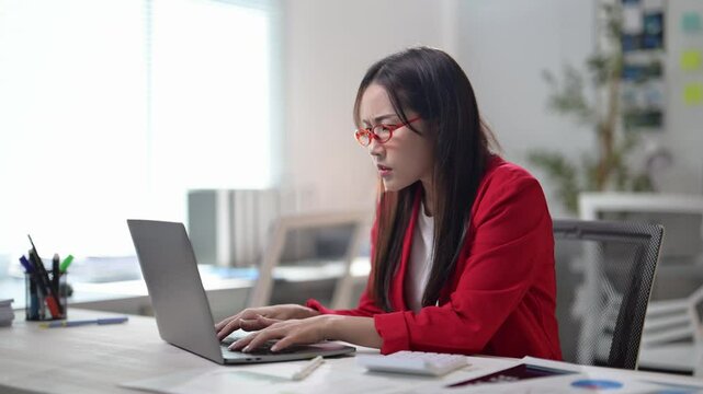 A woman is typing on a laptop computer. She is wearing glasses and a red jacket. There are several pens and a calculator on the desk