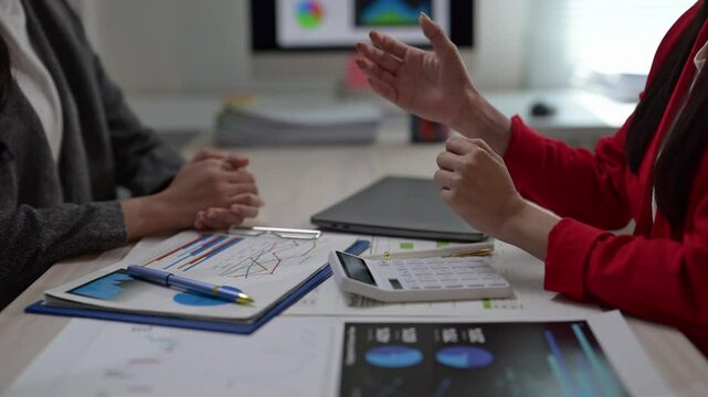 Two women are sitting at a desk with a calculator and a stack of papers. One of the women is wearing a red shirt