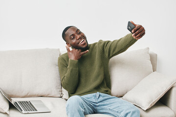 Young Black man smiling and taking a selfie on a couch with a laptop, wearing a green sweater,...