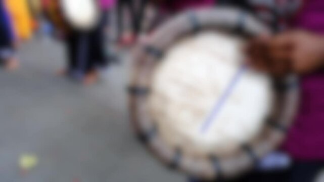 The background features a blurred scene with a large traditional drum and people, suggesting a cultural performance, festival, or ceremonial procession with rhythmic energy.