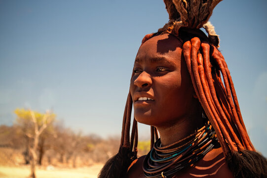 Close-up of an African Himba woman in her village on a sunny day. Her skin and hair are covered with ochre, and traditional jewelry highlights her unique identity
