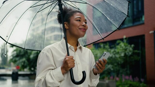 African American woman texting mobile phone smartphone chat online message looking around waiting taxi order meeting female girl businesswoman city outdoors smiling business entrepreneur rain umbrella