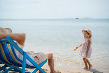 Little girl playing at the beach while a man relaxes in a chair under the sun on a clear day