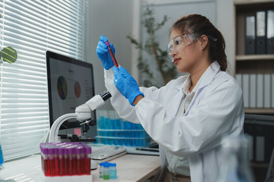 Female scientist wearing lab coat and gloves carefully analyzing blood sample in test tube in modern laboratory with microscope and computer displaying charts - Powered by Adobe
