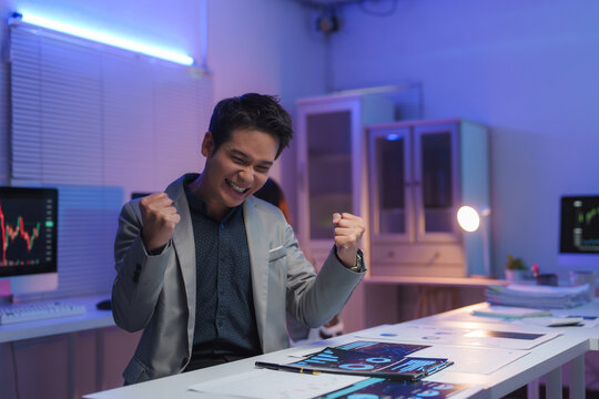 Young businessman raising his fists, feeling excited and happy about achieving business goals, celebrating success in a modern office at night
