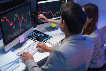 Two financial analysts are working late, examining stock market data on computer screens, with one pointing at a chart with a pen while the other types on a keyboard