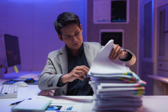 Young businessman reviewing a substantial stack of documents and paperwork while seated at his desk in a modern office, working late into the night, focused on meeting deadlines