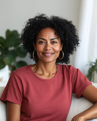 Fototapeta premium a black woman wearing pink t-shirt with no writing or images,sitting comfortably on a light-colored sofa in home,curly hair,large gold hoop earrings,smiling,white interior accents, houseplants