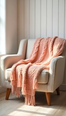 Cozy peach color knitted blanket with fringe on armchair near wall. Home interior with comfy wool throw. Warm textile with braids pattern in sun light. Winter design. House decor.