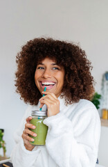 Young mixed race woman drinking healthy green juice in the morning standing in her kitchen at home....