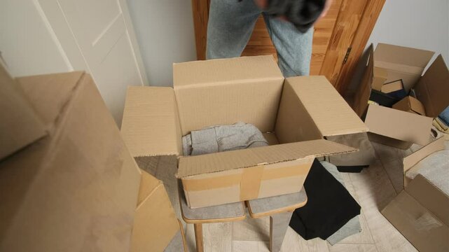 A man places folded clothes into an open cardboard box while preparing for a move. Surrounding him are multiple boxes, scattered clothes, and packing materials in a cluttered room.