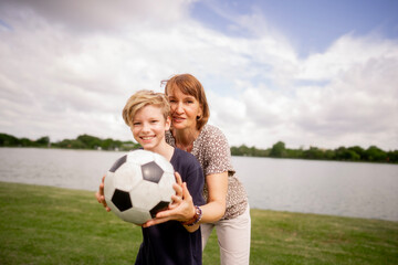 Happy joyful boy hold football to camera with grandmother at summer green grass park. Senior...