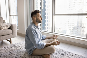 Young Latino man dressed casually sits on lotus position on rug with his eyes closed, fingers in...