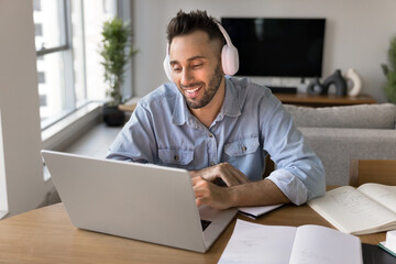 Smiling man sit at desk, wearing headphones working on modern laptop, participating in pleasant online meeting by business or study seated at table with books, textbooks and copybooks. Education, tech
