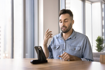 Serious Hispanic man speaking through videocall seated at table, looks at device screen on stand, provide information, professional assistance and advice to client, working remotely using modern app