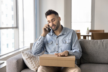 Man sits on couch holding cardboard package while speaking on smartphone, engaged in focused...