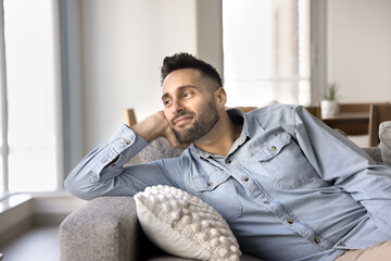 Silent young Hispanic man relaxing leaning on comfortable sofa in contemporary living room, looking...