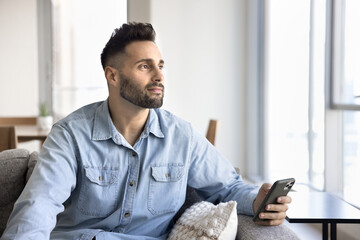Young Hispanic man sit on comfortable couch, holds smartphone, gazing thoughtfully into distance,...