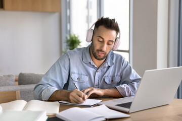 Latin man listening audio course on laptop, studying, preparing university assignment, heap of professional literature, textbooks and copybook spread on table, making notes, write essay, looks focused