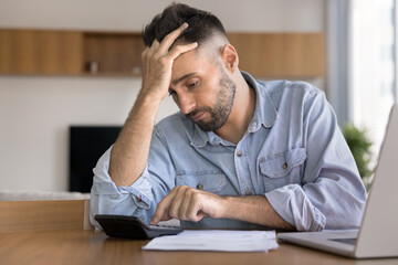 Young 30s Hispanic man feels stressed due to financial issues, while making calculations or budget...