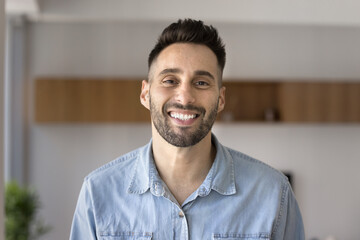 Obraz premium Head shot of handsome young 30s Hispanic man smiling, staring at camera standing alone in cozy domestic room at own or rented apartment. Portrait of friendly millennial Spaniard guy, profile picture