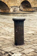 Naklejka premium A worn mooring bollard on a historic cobblestone riverbank, set against the backdrop of an ancient stone bridge. Perfect for themes of history, travel, and urban heritage