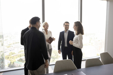 Inspiration from city. Diverse group five businesspeople of all ages genders stand at contemporary designed office meeting room with urban view behind glass walls talk chat enjoy relaxed conversation