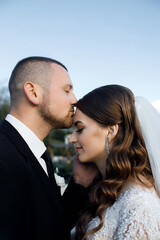 Newlyweds at a wedding photo shoot. The groom in a black suit with a white shirt gently kisses the bride on the forehead.