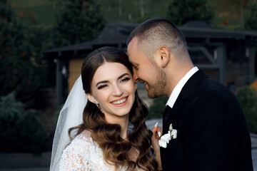 Newlyweds stand on the street, hugging each other. The groom is wearing a black suit with a white...