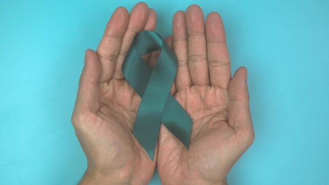 Footage video 4k. Hands holding teal ribbon on blue background.. Ovarian Cancer Awareness, Gynecological, Uterine Cancer, Vulvar Cancer, Panic Disorder, Post Traumatic Stress Disorder (PTSD).
