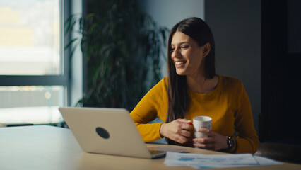Happy young woman smiles while looking at laptop or during a video call, holding a mug in one hand. Concept of remote work, freelancing, business productivity, work-life balance, technology