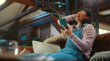 Surprised Japanese woman sitting on couch and reading message with good news on phone, celebrating success, winning using a smartphone. Victory, profit, prize in lottery or online betting.