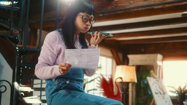 Nervous Japanese woman talking on the phone using speakerphone, complaining and arguing while holding documents at home, high house expenses, and financial business problems.