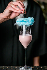 A bartender creates a whimsical cocktail topped with blue cotton candy in a stylish glass. The pink drink contrasts beautifully with the colorful garnish, showcasing creativity.
