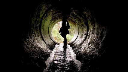 A traveler stands at the end of a dark tunnel, illuminated by natural light. Water trickles underfoot, creating a mysterious but inviting atmosphere for exploration
