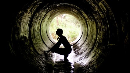 A silhouette of a person crouches inside a circular tunnel, surrounded by lush greenery. This...