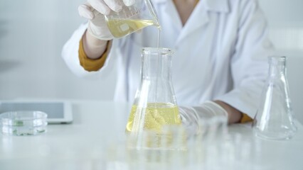 A scientist, wearing a lab coat and white protective gloves, is pouring a yellow oily liquid from one beaker to another in laboratory, close up. Medicine and science concept